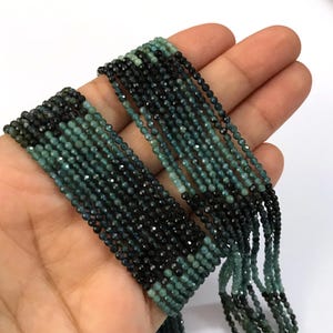May include: A multi-strand necklace featuring small, faceted, round beads in alternating segments of deep black and muted teal green. The beads are strung on multiple parallel lines, creating a textured, layered appearance with a consistent color pattern.