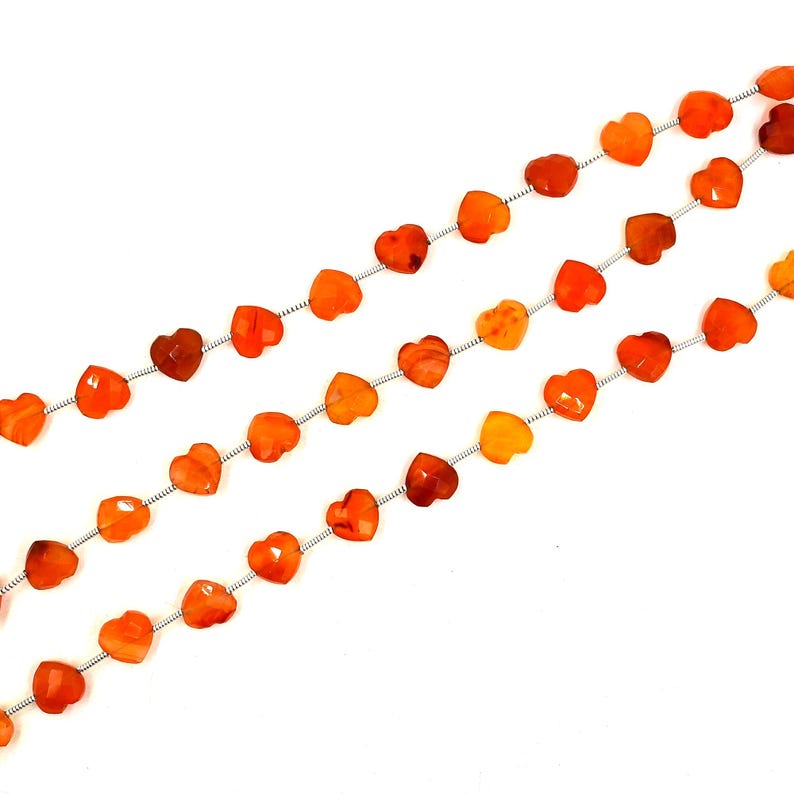 May include: A strand of faceted orange carnelian beads shaped like small hearts. The beads are strung on a thin silver-colored wire, arranged in three parallel lines across the frame, showcasing a consistent vibrant orange hue throughout.