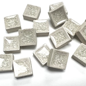 May include: A collection of 13 small, white, square ceramic tiles with a textured surface. The tiles are arranged in a random pattern on a white background.