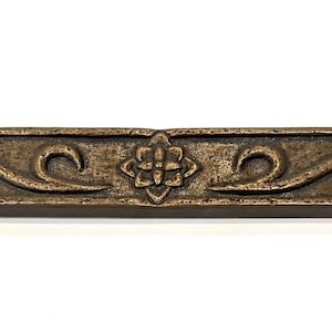 May include: A decorative tile with a brown and gold finish. The tile features a floral design with intricate swirls and a central flower.