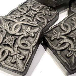 May include: Close-up of several gray, rectangular decorative tiles with an ornate, repeating pattern. The design features intertwined loops, floral elements, and a central starburst. The tiles are arranged on a white surface.