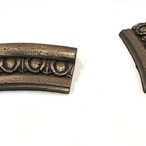 May include: Two curved pieces of decorative molding with a textured, metallic bronze finish. The molding features a repeating pattern of raised circles and leaves.