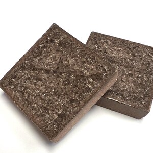 May include: Two square, brown coasters with a textured, crystalline surface. The coasters are made of a dark brown material and have a slightly raised edge. They are set against a white background.
