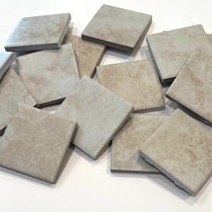 May include: A collection of square tiles with a textured, light beige surface and dark brown edges. The tiles are scattered, showcasing their individual details and variations in color. These tiles are suitable for home improvement projects.