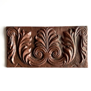 Puede incluir: Un azulejo decorativo con un diseño en relieve de hojas y espirales estilizadas. El azulejo está hecho de un material marrón y tiene un aspecto rústico y envejecido.