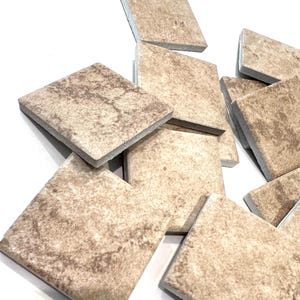 May include: A collection of beige square tiles with a textured, marbled appearance. The tiles are arranged in a slightly scattered pile, showcasing their varying patterns and shades of brown and tan. The edges are clean and the overall aesthetic is neutral.