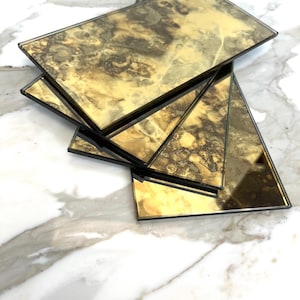 May include: A stack of rectangular, antique-style glass tiles with a distressed gold and brown finish. The tiles have black edges and are arranged in a fan-like pattern, showcasing their unique, aged appearance. The background is a white marble surface.