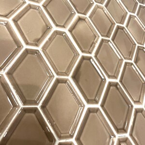 May include: A close-up of a wall tiled with hexagonal tiles. The tiles are a light brown color with white grout lines. The tiles have a slightly raised edge around the perimeter.