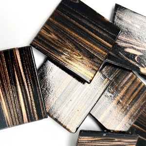 May include: A set of eight square coasters with a black and gold woodgrain pattern. The coasters are made of a smooth, glossy material.