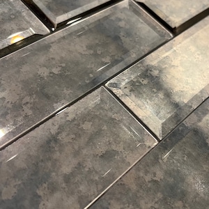 May include: Close-up of a gray and brown glass tile with a beveled edge. The tile has a textured surface that resembles aged metal.