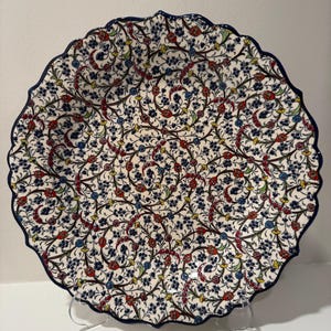 May include: A decorative ceramic plate with a white background and a detailed floral pattern in red, blue, and yellow. The plate has a scalloped edge with a dark blue border. The design features intricate vines and flowers.