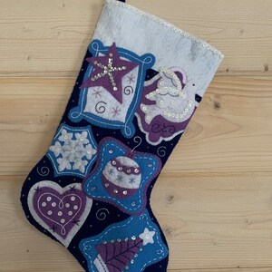 May include: A blue felt Christmas stocking with a white cuff. The stocking is decorated with purple, blue, and white felt ornaments, including a star, a heart, a snowflake, a Christmas tree, and a bell. The ornaments are embellished with sequins.