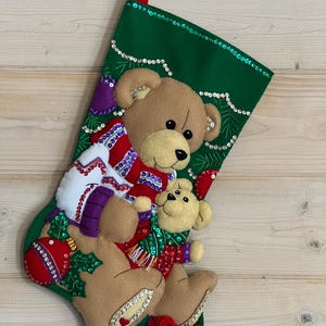 May include: A green Christmas stocking with a red loop, decorated with felt teddy bears, ornaments, and a gift box. The stocking is embellished with sequins and has a festive holiday theme.