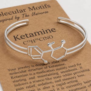 May include: A silver-toned metal cuff bracelet featuring a wire-frame design of the Ketamine molecular structure. The bracelet is displayed on a rectangular brown card printed with the text Molecular Motifs, Ketamine C13H16ClNO, and descriptive information about the chemical compound.