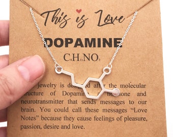 May include: A silver necklace with a pendant shaped like the chemical structure of dopamine. The pendant is on a brown card with the text "This is Love" and "Dopamine CHNO".