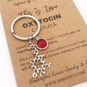 May include: A silver-toned metal keychain featuring a molecular structure charm of oxytocin and a small circular red charm. The keychain is attached to a tan card printed with the text Molecular Motifs, This is Love, OXYTOCIN, and chemical formula.