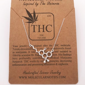 Peut inclure: Un collier en argent avec un pendentif en forme de structure moléculaire de THC. Le pendentif est sur une carte brune avec le texte "Molecular Motifs Inspired by The Universe" et "THC 710.000".