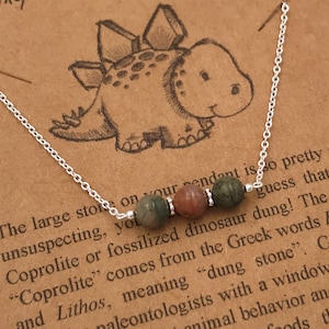 May include: A silver necklace with a charm of a cartoon dinosaur and three green and brown gemstone beads. The necklace comes with a card that reads "The large stone on your pendant is so pretty and unsuspecting, you might guess that it's Coprolite or fossilized dinosaur dung! The word "Coprolite" comes from the Greek words Kopros and Lithos, meaning "dung stone". Coprolite provides paleontologists with a window into the dynamic of ancient animal behavior and biodiversity, giving us insights into what foods the dinosaurs had for dinner."