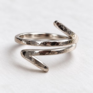 May include: A silver-toned bypass ring featuring a hammered texture across the entire surface. The band wraps around the finger with two open, pointed ends that overlap slightly, creating a continuous, angular, and reflective metallic design.