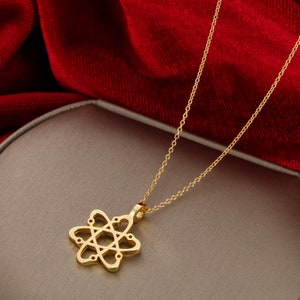 May include: A gold chain necklace with a pendant shaped like an atom with hearts for the electrons. The necklace is on a light brown surface.
