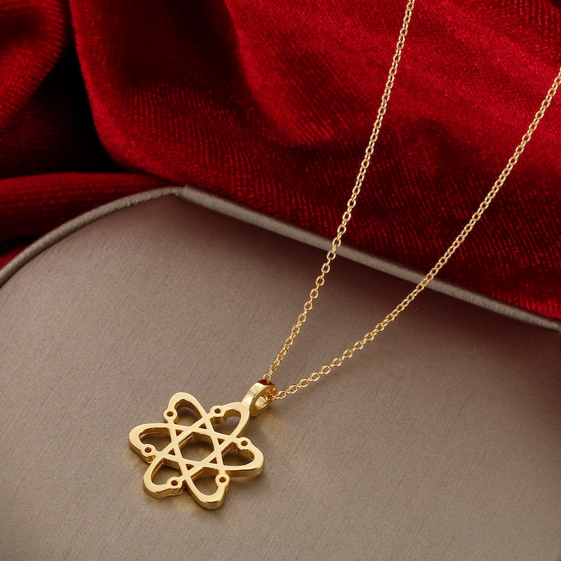May include: A gold chain necklace with a pendant shaped like an atom with hearts for the electrons. The necklace is on a light brown surface.