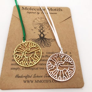 May include: Two gold-coloured tree of life pendants on white and green ribbons. The pendants are circular and feature a detailed design of a tree of life. The background is a brown card with the text "Molecular Motifs Inspired by The Universe" and "Handcrafted Science Jewellery & Gifts" and the website "www.mmmotifs.com".