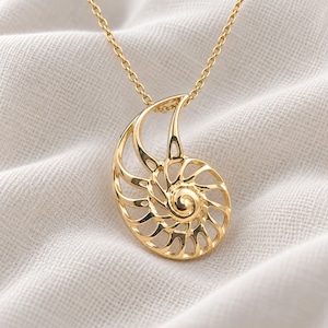 May include: A gold-toned necklace featuring a pendant shaped like an open-work nautilus shell. The pendant has a polished, reflective surface and is suspended from a delicate matching gold-toned chain with small, uniform circular links.