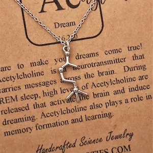 May include: A silver necklace with a pendant shaped like the chemical structure of acetylcholine, a neurotransmitter associated with dreams and memory. The card reads "Acetylcholine is a neurotransmitter that carries messages across the brain. During REM sleep, high levels of Acetylcholine are released that activate the brain and induce dreaming. Acetylcholine also plays a role in memory formation and learning." The card also reads "Dream" and "Handcrafted Science Jewelry www.mmotifs.com".
