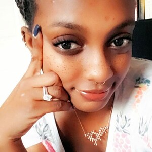 May include: A close-up of a person's face with a nose ring and a gold necklace with a geometric design. The person is wearing a white shirt with a floral pattern.