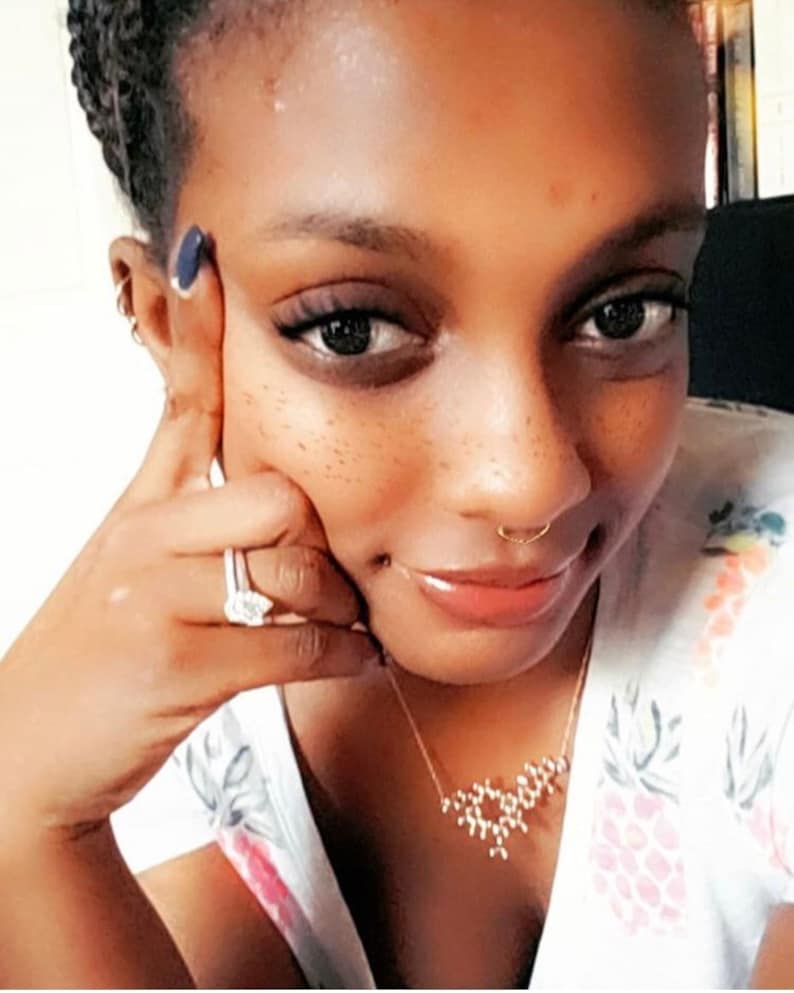 May include: A close-up of a person's face with a nose ring and a gold necklace with a geometric design. The person is wearing a white shirt with a floral pattern.
