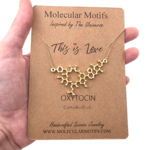 May include: A gold necklace with a pendant shaped like the molecular structure of oxytocin. The pendant is on a brown card with the text "Molecular Motifs Inspired by The Universe This is Love OXYTOCIN C43H66N12O12S2 Handcrafted Science Jewelry WWW.MOLECULARMOTIFS.COM".