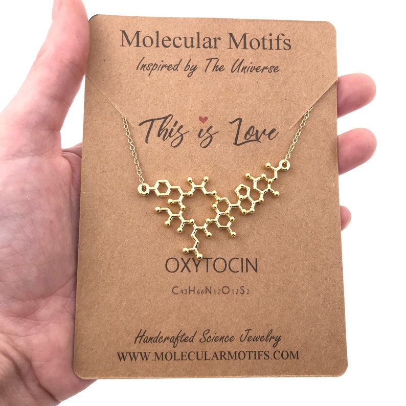 May include: A gold necklace with a pendant shaped like the molecular structure of oxytocin. The pendant is on a brown card with the text "Molecular Motifs Inspired by The Universe This is Love OXYTOCIN C43H66N12O12S2 Handcrafted Science Jewelry WWW.MOLECULARMOTIFS.COM".