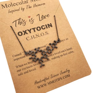 May include: A black necklace with a pendant shaped like the molecular structure of oxytocin. The pendant is on a brown card with the text "This is Love" and "OXYTOCIN C43H66N12O12S2" in white. The card also includes text about oxytocin being the "love hormone" and its role in bonding.