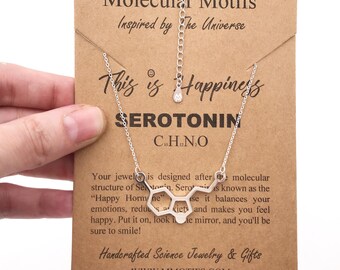 May include: A silver-toned necklace featuring a serotonin molecular structure pendant on a delicate chain. The necklace is displayed on a rectangular brown card printed with the text Molecular Motifs, This is Happiness, SEROTONIN, and C10H12N2O.