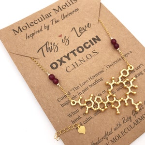May include: A gold necklace with a pendant shaped like the molecular structure of oxytocin, the "love hormone". The necklace is on a brown card with the text "This is Love OXYTOCIN C8H13N2O5S" and "Handcrafted with Ruby Garnet Gemstones MOLECULARMOTIFS.COM".