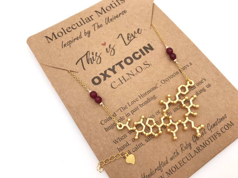 May include: A gold necklace with a pendant shaped like the molecular structure of oxytocin, the "love hormone". The necklace is on a brown card with the text "This is Love OXYTOCIN C8H13N2O5S" and "Handcrafted with Ruby Garnet Gemstones MOLECULARMOTIFS.COM".
