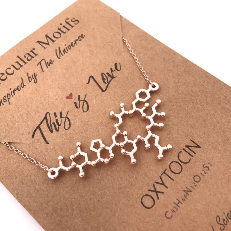 May include: Rose gold necklace with a pendant shaped like the molecular structure of oxytocin. The pendant is on a brown card with the text "This is Love" and "Molecular Motifs inspired by The Universe". The card also includes the chemical formula for oxytocin: C43H66N12O12S2.