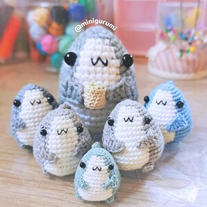 Pu&ograve; includere: Un gruppo di sei peluche di squalo all'uncinetto in varie tonalit&agrave; di blu e grigio. I peluche hanno occhi neri e sorrisi bianchi. I peluche sono seduti su una superficie marrone chiaro. Il testo '@minigurumi' &egrave; visibile sullo sfondo.