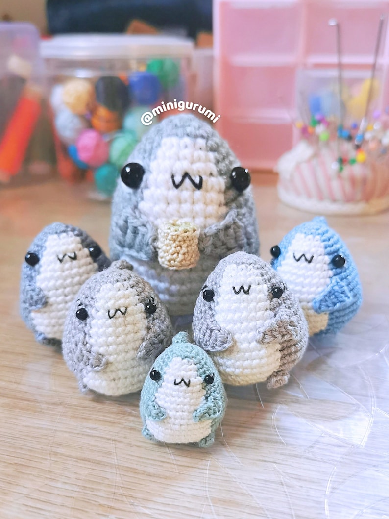 Pu&ograve; includere: Un gruppo di sei peluche di squalo all'uncinetto in varie tonalit&agrave; di blu e grigio. I peluche hanno occhi neri e sorrisi bianchi. I peluche sono seduti su una superficie marrone chiaro. Il testo '@minigurumi' &egrave; visibile sullo sfondo.