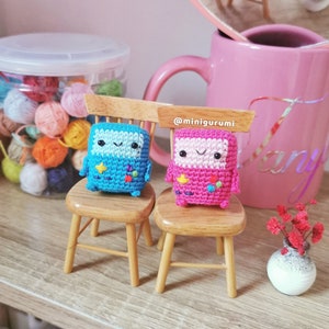 May include: Two small crocheted figures, one blue and one pink, are sitting on miniature wooden chairs. The figures have smiling faces and pixelated designs. The chairs are on a wooden shelf with a jar of yarn balls in the background. The text '@minigurumi' is visible on the shelf.