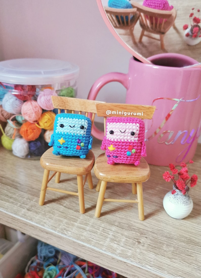 May include: Two small crocheted figures, one blue and one pink, are sitting on miniature wooden chairs. The figures have smiling faces and pixelated designs. The chairs are on a wooden shelf with a jar of yarn balls in the background. The text '@minigurumi' is visible on the shelf.