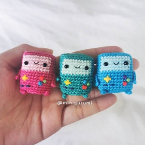 May include: Three crocheted Game Boy characters, one pink, one teal, and one blue, with smiling faces and pixelated details.