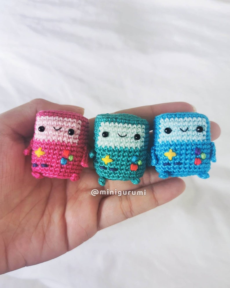 May include: Three crocheted Game Boy characters, one pink, one teal, and one blue, with smiling faces and pixelated details.