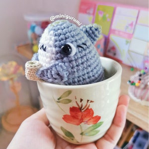 Pu&ograve; includere: Un animale di uncinetto grigio e bianco con grandi occhi &egrave; seduto in una piccola tazza da t&egrave; con motivo floreale. La tazza &egrave; tenuta in una mano. Il testo '@minigurumi' &egrave; visibile sullo sfondo.