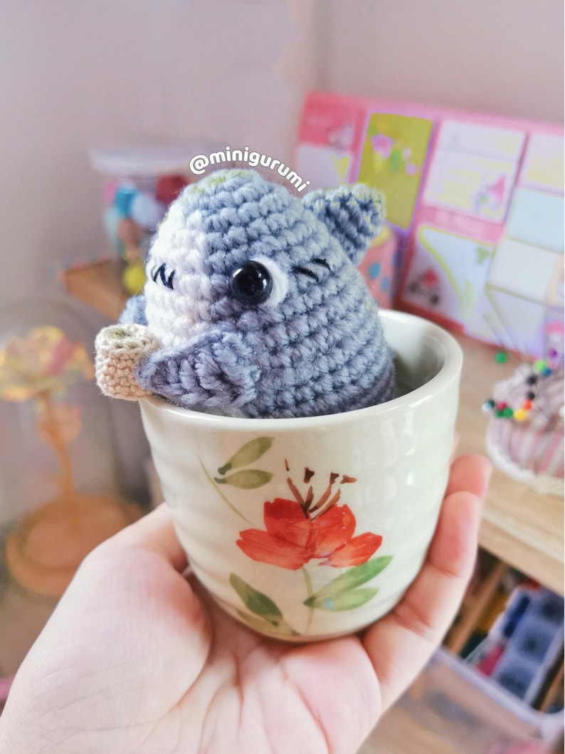 Pu&ograve; includere: Un animale di uncinetto grigio e bianco con grandi occhi &egrave; seduto in una piccola tazza da t&egrave; con motivo floreale. La tazza &egrave; tenuta in una mano. Il testo '@minigurumi' &egrave; visibile sullo sfondo.