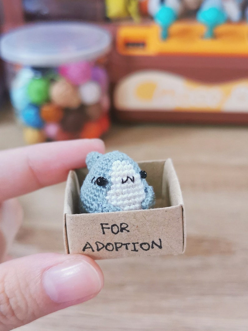 Pu&ograve; includere: Un piccolo animale di uncinetto grigio con occhi neri e un naso bianco &egrave; seduto in una scatola di cartone. La scatola ha le parole "FOR ADOPTION" scritte su di essa.