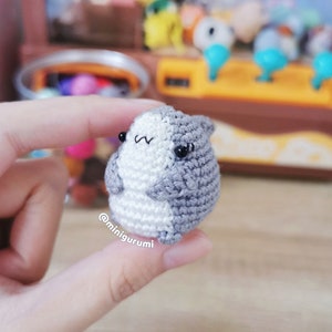 Pu&ograve; includere: Un piccolo animale amigurumi di uncinetto grigio e bianco con occhi neri. L'animale &egrave; tenuto in una mano.