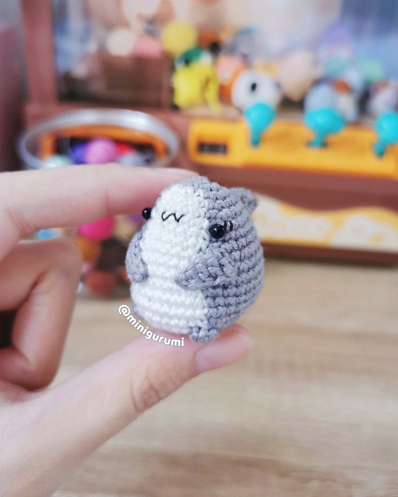 Pu&ograve; includere: Un piccolo animale amigurumi di uncinetto grigio e bianco con occhi neri. L'animale &egrave; tenuto in una mano.