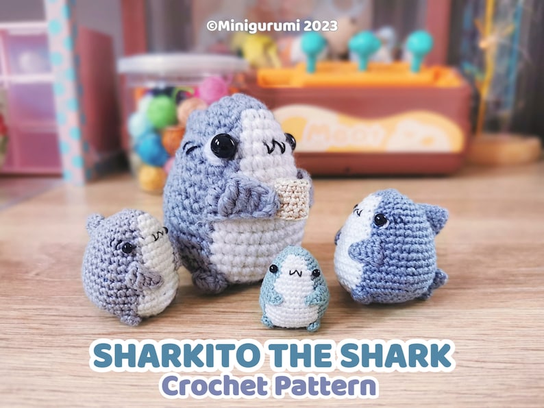 Pu&ograve; includere: Quattro peluche di squalo all'uncinetto di diverse dimensioni. Lo squalo pi&ugrave; grande &egrave; grigio e bianco con occhi neri e tiene un piccolo rettangolo all'uncinetto. Gli altri tre squali sono tutti blu e bianchi con occhi neri. I peluche sono seduti su una superficie di legno. "Sharkito the Shark Crochet Pattern" &egrave; scritto in lettere blu sotto i peluche.