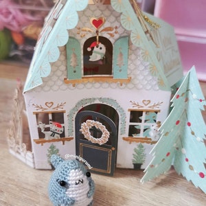 Pu&ograve; includere: Una piccola statuina di pinguino in crochet blu e bianco si trova davanti a una casa di Natale in carta con una ghirlanda sulla porta e un albero di Natale a destra. La casa &egrave; decorata con glitter e ha un piccolo uccello in una finestra.